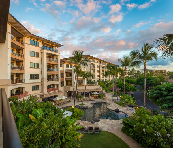 The image shows a resort with multi-story buildings, a pool area, palm trees, lush greenery, and a scenic sky with clouds during sunset.