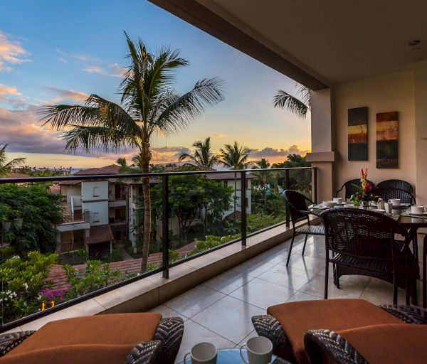 A balcony with outdoor seating, a dining table set for a meal, and a picturesque view of palm trees and buildings at sunset, creating a serene ambiance.