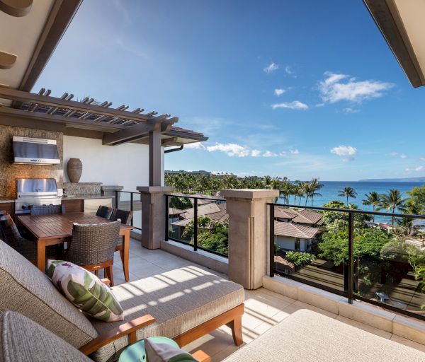 A balcony with lounge chairs, a dining area, and a grill overlooks a scenic coastal view with palm trees, rooftops, and a clear blue sky.
