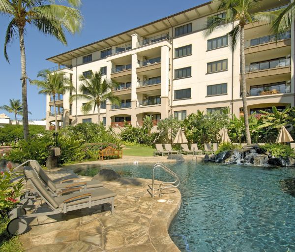 This image shows a resort with a swimming pool, lounge chairs, tropical plants, and a multi-story building in the background.