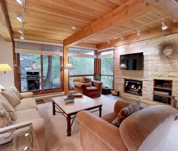 A cozy living room with wooden beams, large windows, a stone fireplace, TV, and comfortable seating, with nature visible outside the windows.