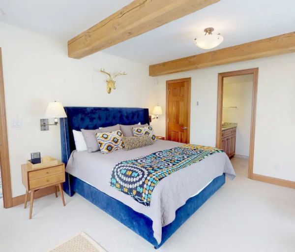 A cozy bedroom with a blue bed, patterned bedding, wooden beams, and a flat-screen TV. An adjacent room with large windows is visible through the doorway.