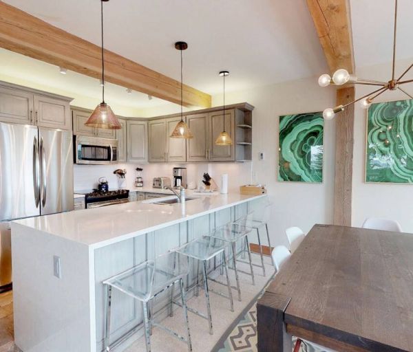 This image shows a modern kitchen with stainless steel appliances, an island with clear barstools, a wooden dining table, and contemporary light fixtures.