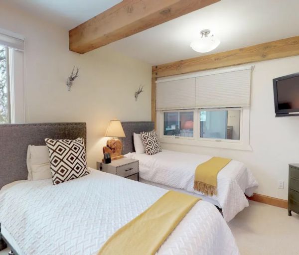 A cozy bedroom with two single beds, patterned pillows, a wall-mounted TV, window, lamp, and a small chest of drawers.