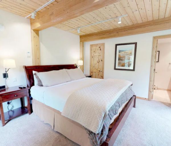 This image shows a cozy bedroom with wooden paneling, a large bed, nightstands, and a chair. A doorway leads to a bathroom with a wooden door.