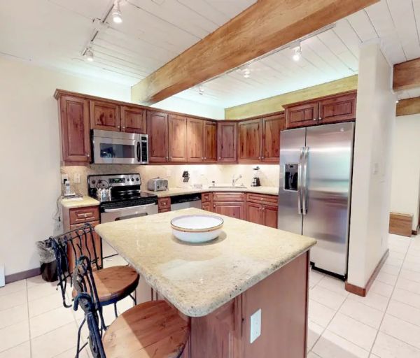 A modern kitchen features wooden cabinets, stainless steel appliances, an island with seating, and an open doorway leading to a bedroom.
