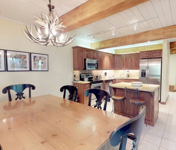 A kitchen and dining area with wooden furniture, antler chandelier, stainless steel appliances, and framed art on the wall, ending the sentence.