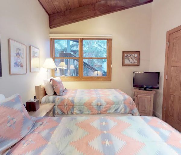A cozy bedroom with two beds in soft geometric-patterned covers, wooden furnishings, wall art, a TV, and a window showing an outdoor view.