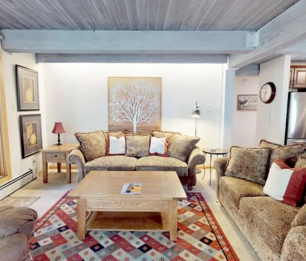 A cozy living room with a mix of cushioned and wooden furniture, artwork on the walls, and an adjacent kitchen with modern appliances.