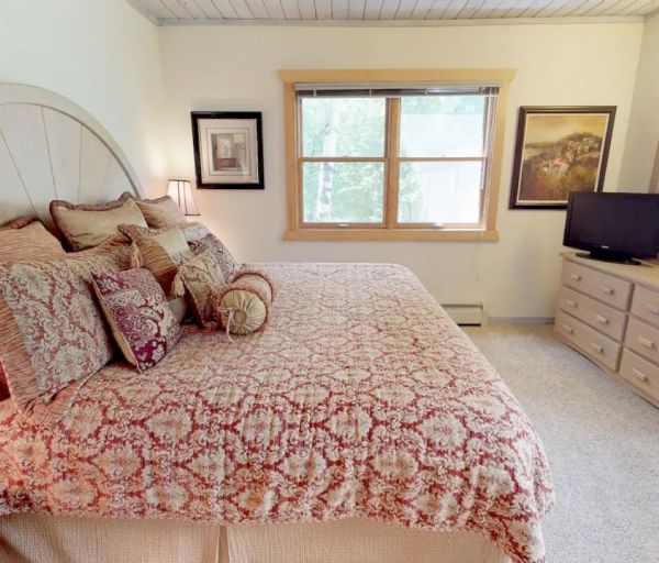 A cozy bedroom with a large bed, decorative pillows, a dresser with a mirror, a flat-screen TV, and framed artworks on the walls.