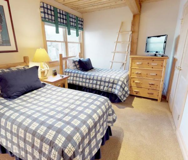 A cozy bedroom with two single beds, plaid bedding, a wooden dresser with a TV, a window, and Native American-themed decor on the wall.