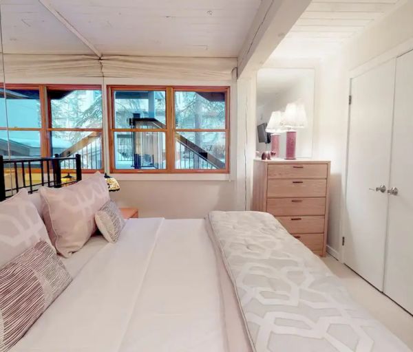 A cozy bedroom with a bed, pillows, side table, dresser, lamp, large window, closet, and a wall-mounted TV.