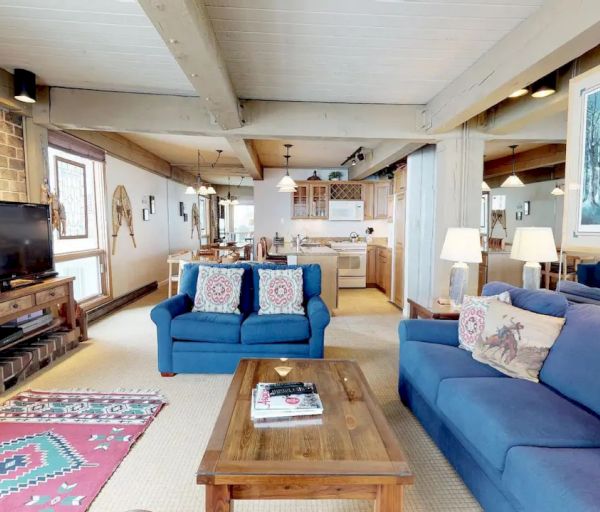 A cozy living room with blue sofas, a wooden coffee table, a TV, a fireplace, and an open kitchen in the background, with rustic decor.