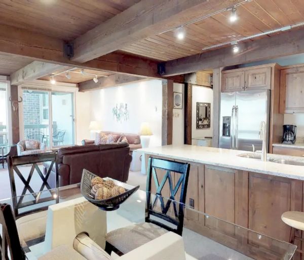 A cozy open-plan living area with wooden ceilings, including a kitchen with a breakfast bar, dining area, and lounge with large windows.