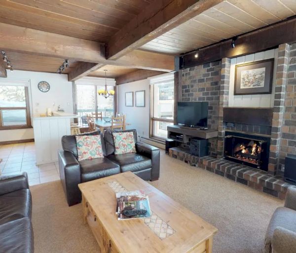 A cozy living room with leather sofas, a wooden coffee table, a brick fireplace, a TV, and visible kitchen and dining areas through an open layout.