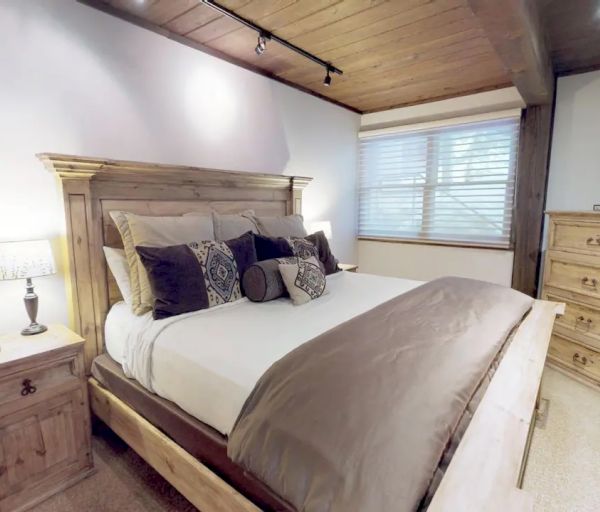A cozy bedroom with a wooden bed, nightstand with a lamp, dresser with a TV, carpeted floor, and a window with blinds ending the sentence.