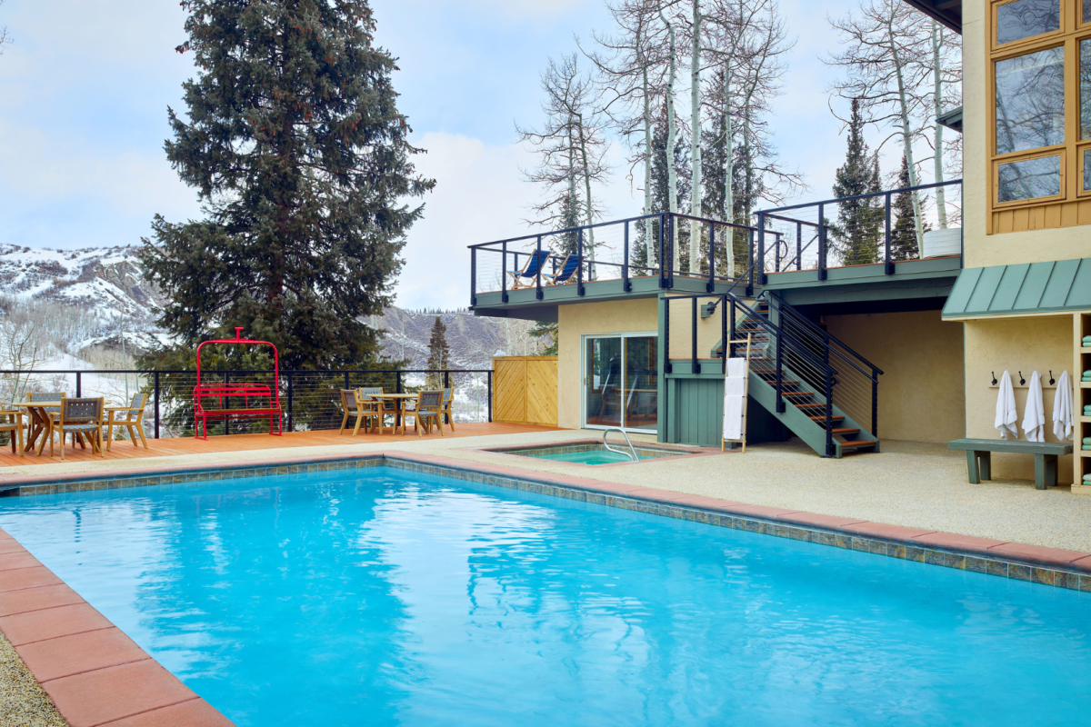 The image shows a serene outdoor setting with a swimming pool, wooden patio furniture, a red bench, trees, and a surrounding mountainous landscape.