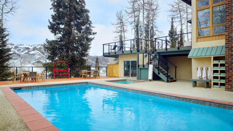 The image shows a serene outdoor setting with a swimming pool, wooden patio furniture, a red bench, trees, and a surrounding mountainous landscape.