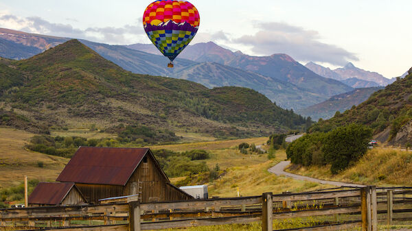 A colorful hot air balloon floats over a rural landscape with a barn, hills, and a winding road in the background.