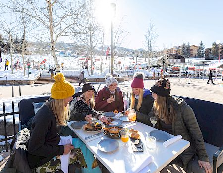 A group of people in winter attire are sitting outdoors around a table eating and drinking, with a snowy landscape and skiing area in the background.