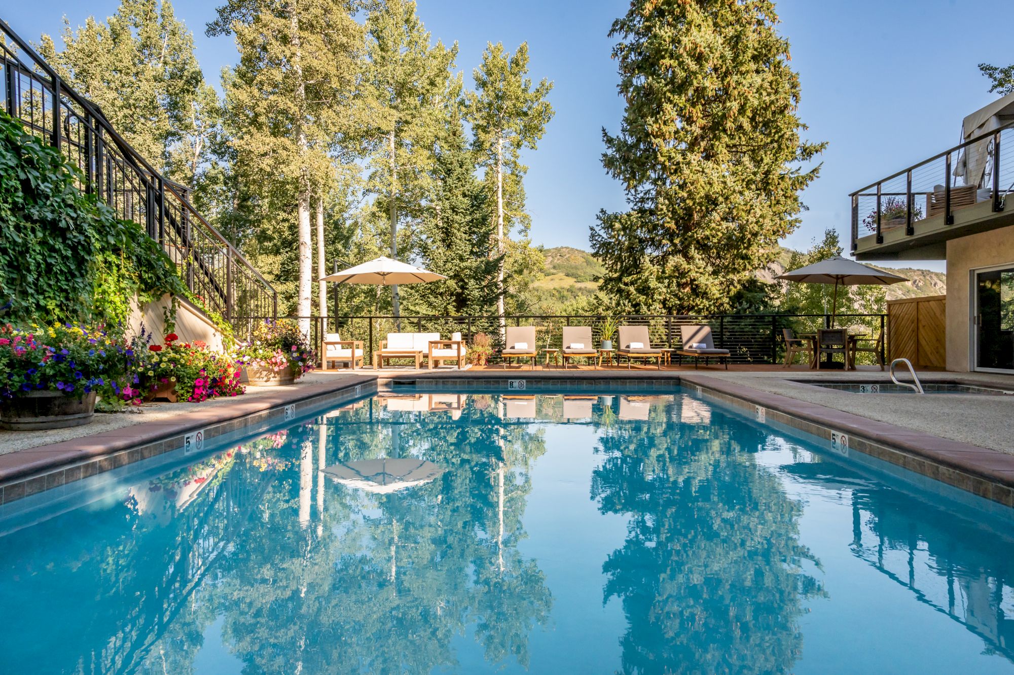 A serene outdoor pool area surrounded by trees and lounge chairs with umbrellas, reflecting blue skies and greenery in the calm water.