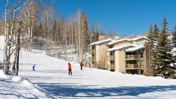 The image shows a snowy landscape with skiers, tall trees, and nearby accommodations under a clear blue sky. Winter vibes abound.