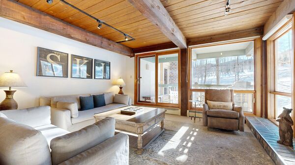 A cozy living room with a beige sofa set, neutral armchairs, wooden ceiling beams, large windows, and a glass door leading outside, bathed in natural light.