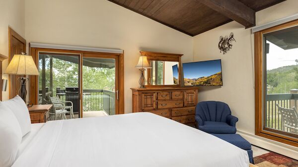The image shows a cozy bedroom with a large bed, wooden furniture, a TV, and a view of a deck and greenery outside.
