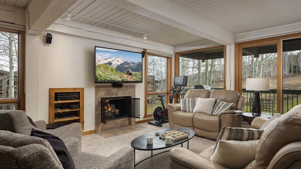 Cozy living room with a stone fireplace, a flat‑screen TV above, wood trim, large windows, and multiple beige fabric sofas arranged around a glass coffee table.