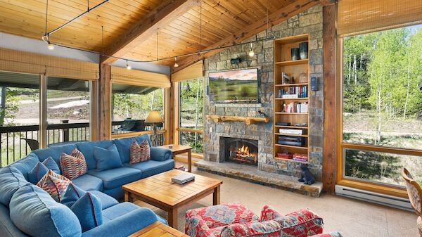 A cozy living room with a blue couch, colorful chairs, stone fireplace, bookshelves, wooden ceiling, and large windows showcasing a forest view.