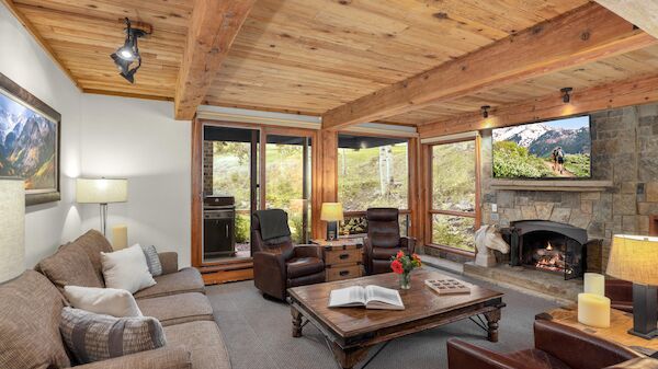 The image shows a cozy living room with wooden beams, a stone fireplace, comfortable seating, and large windows showcasing nature.