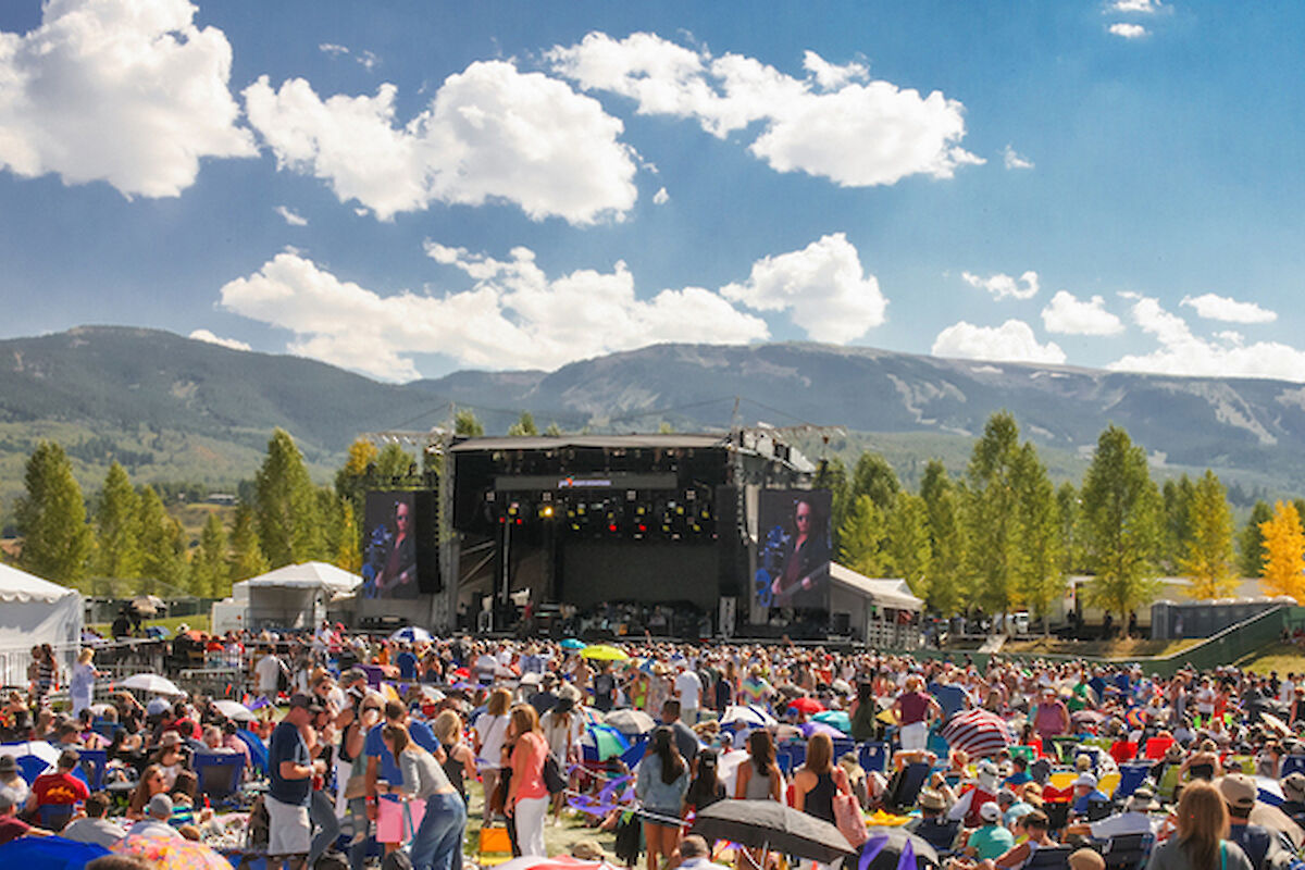The image shows a large crowd at an outdoor music festival, with a stage set against a backdrop of mountains and blue skies.