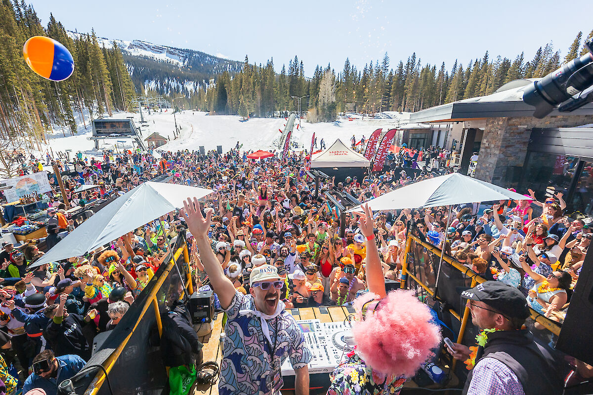 A lively outdoor party scene on a snowy mountain, with a DJ performing and a vibrant crowd celebrating in colorful attire.