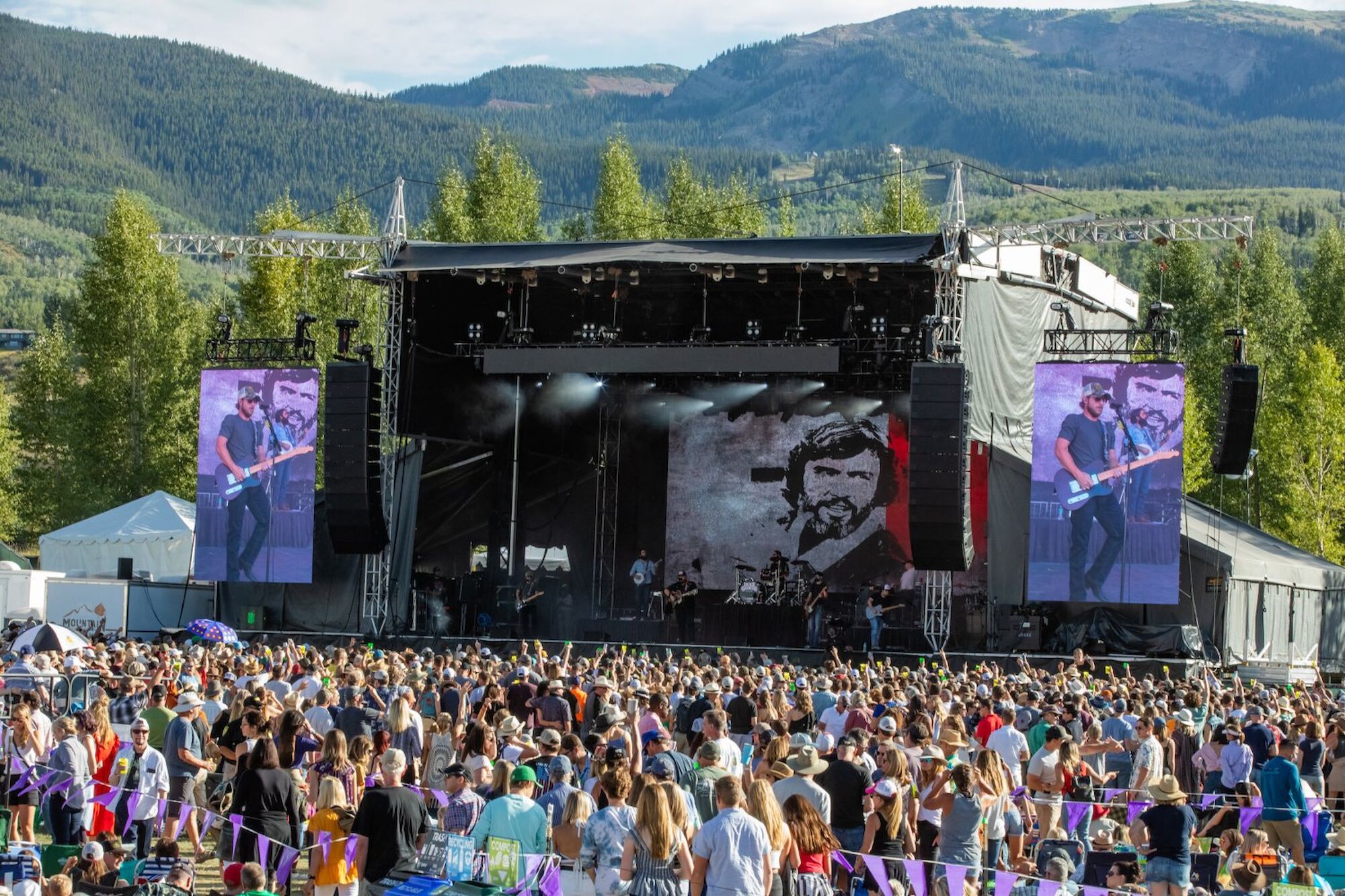The image shows a large outdoor concert stage with a crowd gathered in front, set against a scenic backdrop of mountains.