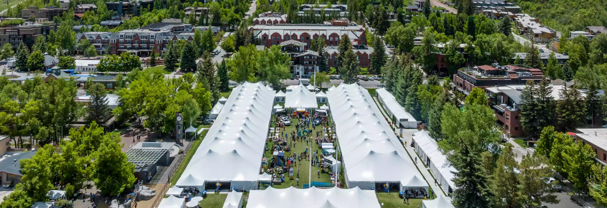 The image shows a large outdoor event area with white tents and green trees, likely a festival or market. It has a vibrant atmosphere.