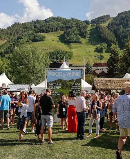 A lively outdoor festival scene with tents and crowds, set against a scenic mountain backdrop. People enjoy food and wine.