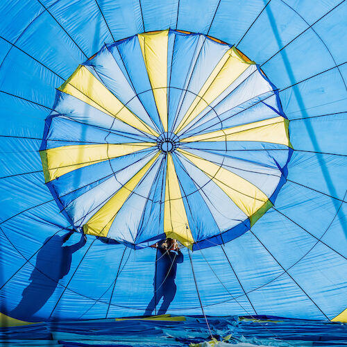 The image shows two people handling a large, bright blue and yellow parachute, shaping it as they prepare for flight.