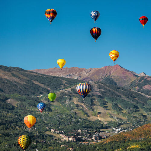 The image shows colorful hot air balloons floating over a mountainous landscape under a clear blue sky. It's a vibrant scene.
