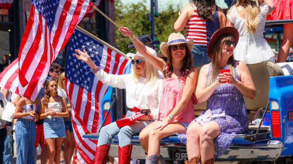 A festive parade scene with women wearing cowboy hats, waving flags, and sitting on a truck bed, while kids and spectators cheer nearby.