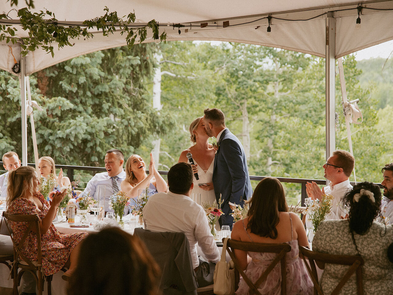 A couple kisses at a wedding reception under a canopy, guests seated around tables, greenery outside, soft festive vibes.