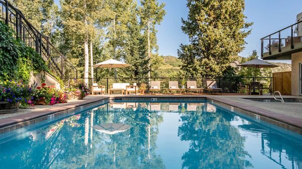 A sunny pool area with clear blue water, lounge chairs, umbrellas, and surrounding greenery reflecting in the water, at a cozy resort.