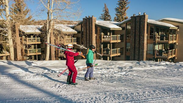 Two people ski together in a snowy courtyard with brown brick buildings and leafless trees in the background.