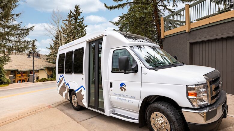 A white passenger van parked on a driveway near a building, with open side door and blue logo on the side, trees in the background, sunny day.