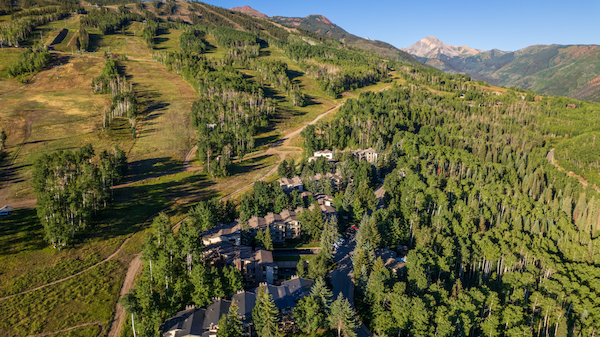 A hillside mountain village with trees, winding roads, and scattered houses nestled in a green valley under clear blue skies.