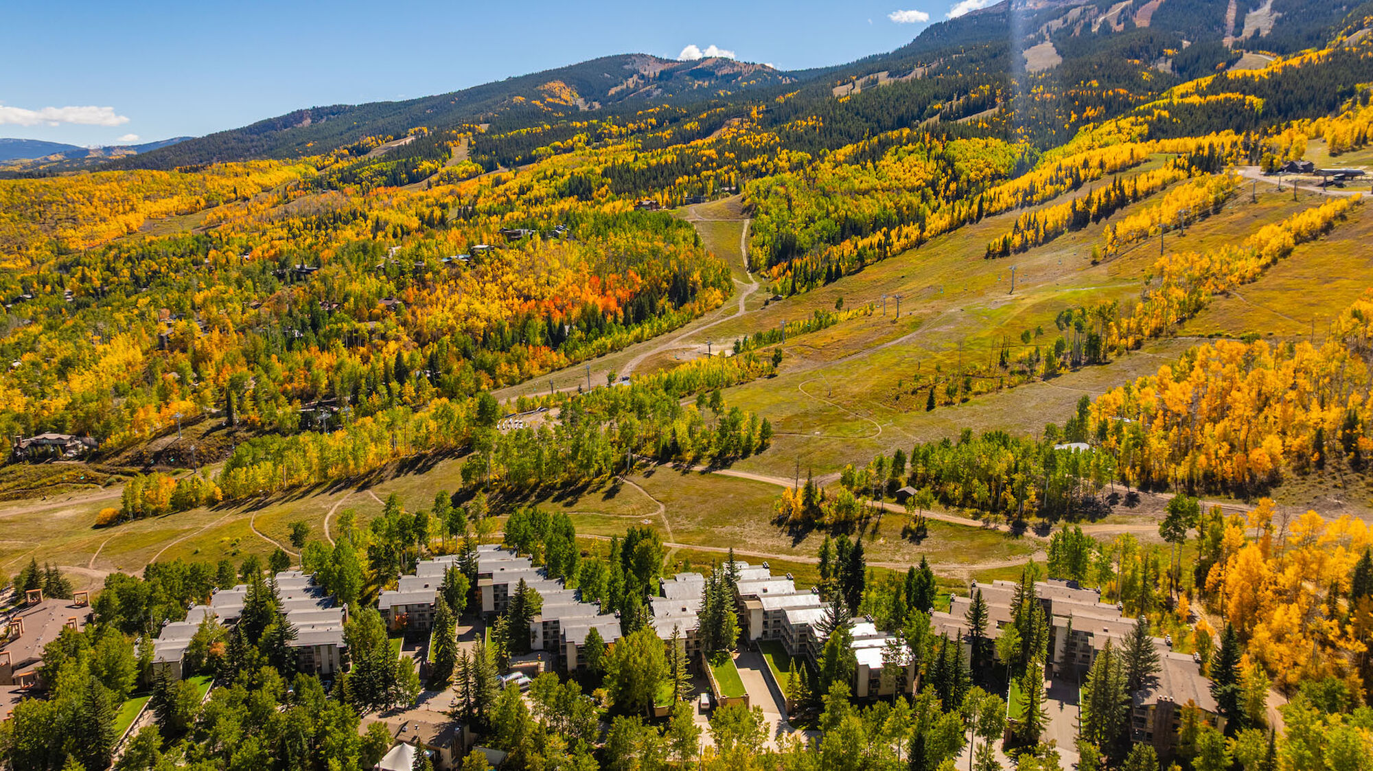 A sweeping mountain valley with vibrant autumn foliage, a small resort or village clustered near pine trees, and winding trails across golden hills.
