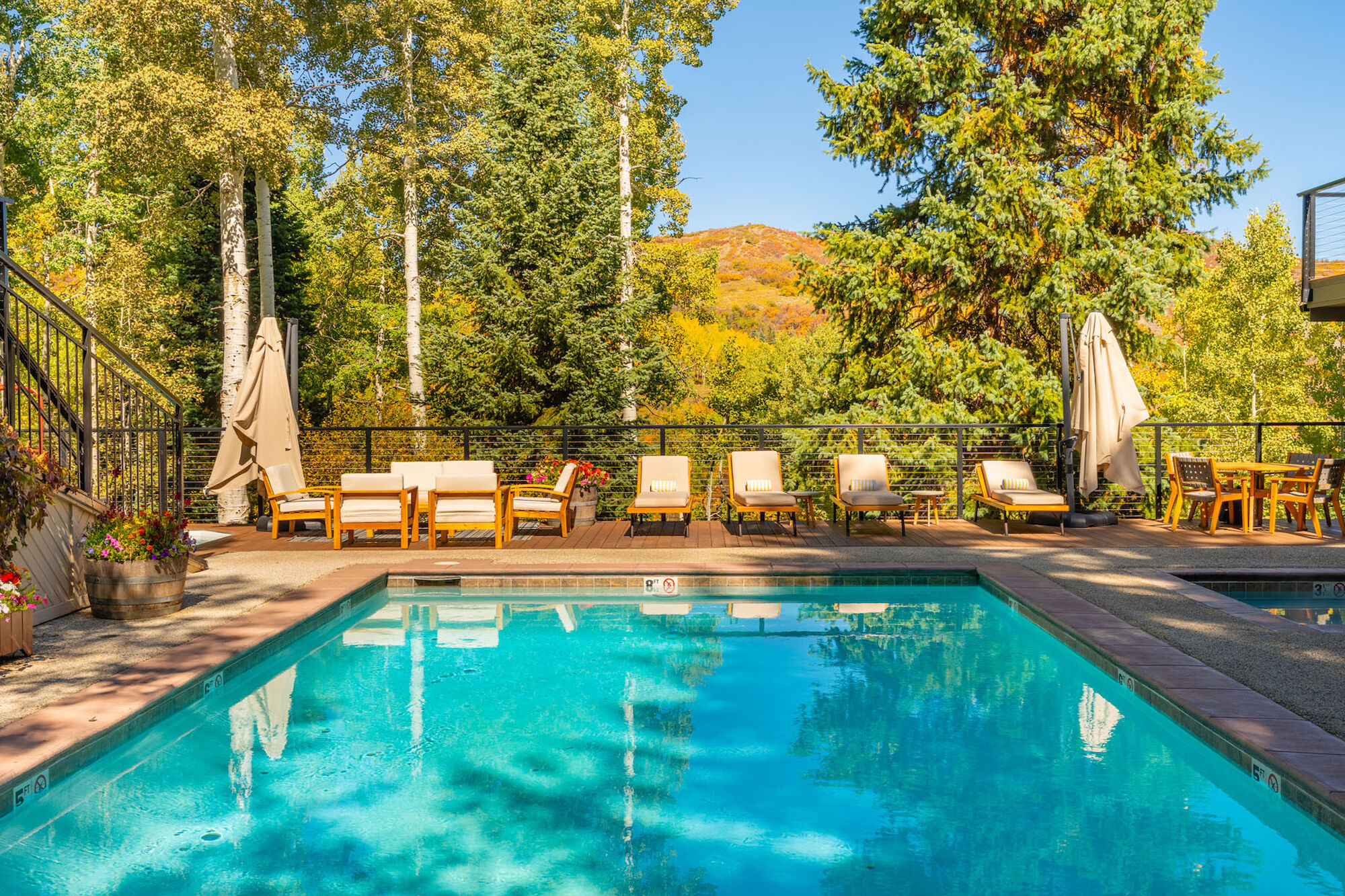 A bright outdoor pool surrounded by lounge chairs, umbrellas, and trees with hills in the background, sunny and inviting.