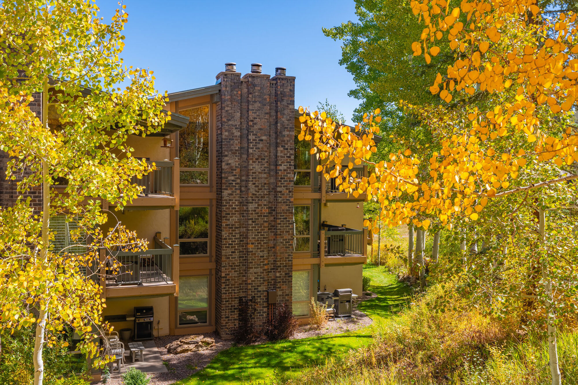 A two-story brick apartment building nestled among vibrant fall trees with yellow leaves and green grass, balconies on each side, bright daylight.